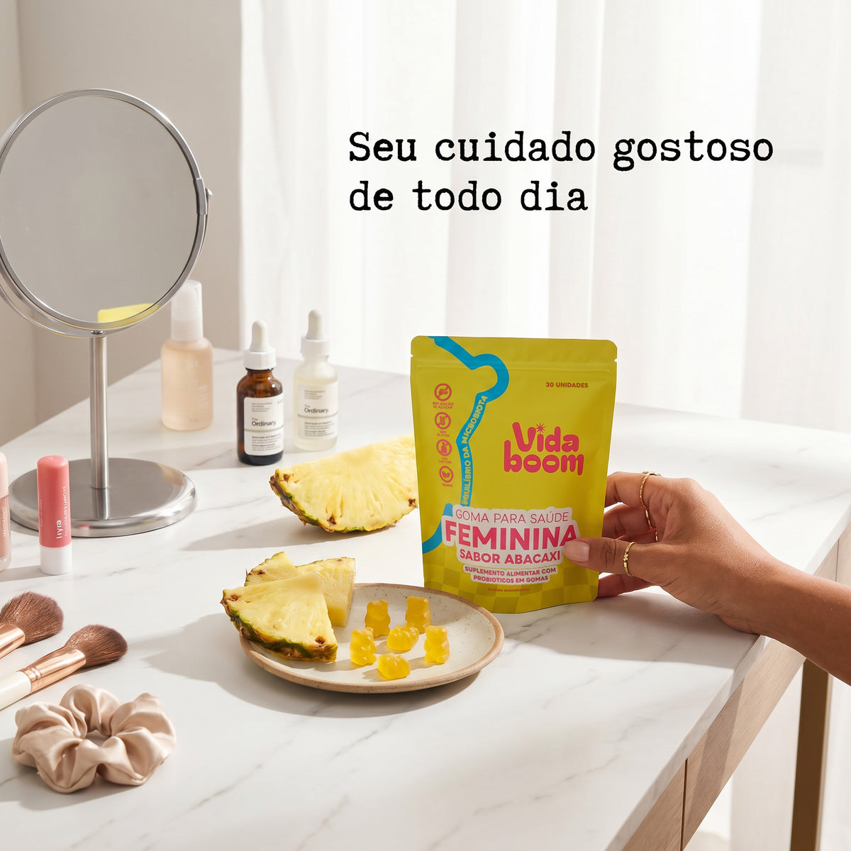 Mão segurando a Goma para Saúde Feminina sabor abacaxi na penteadeira, com gomas de ursinho e fatias de abacaxi
