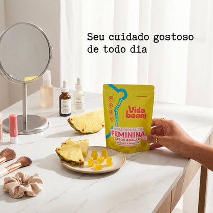 Mão segurando a Goma para Saúde Feminina sabor abacaxi na penteadeira, com gomas de ursinho e fatias de abacaxi